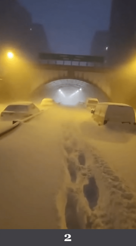 Verschneite Tunnelzufahrt mit parkenden Fahrzeugen und tiefem Schneefall. Beschreibung: Mehrere Fahrzeuge stehen in einer schneebedeckten Straße vor einer Tunnelöffnung. Schneespuren verlaufen in Richtung Tunnel, Beleuchtung wirkt gleichmäßig verteilt. Könnte das real sein? Nein. Die Schneeverteilung an Fahrzeugen und Fahrbahn ist auffallend symmetrisch und wirkt wie eine Texturauflage, nicht wie windgetriebene Ablagerung.